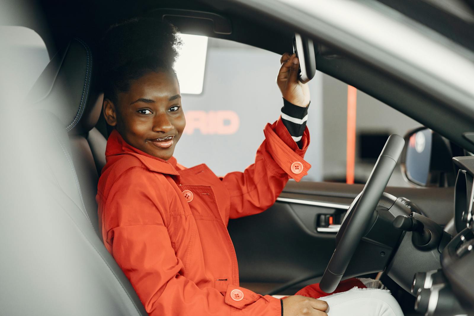 A woman in a red jacket sits in a car, adjusting the rearview mirror and smiling at the camera.